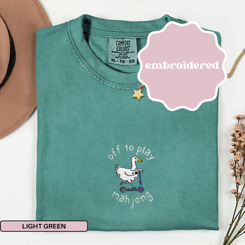 May include: Light green Comfort Colors t-shirt with an embroidered design. The design features a white duck riding a blue scooter with the words "off to play mahjong". The shirt has a tag that says XL-TG-EG.