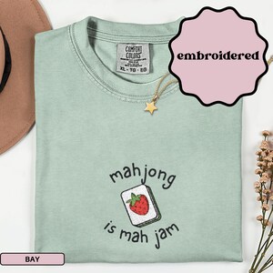May include: A light green t-shirt with the text "mahjong is mah jam" and an embroidered strawberry tile design. The shirt is a Comfort Colors brand, size XL-TG-EG. A gold star necklace is also visible.