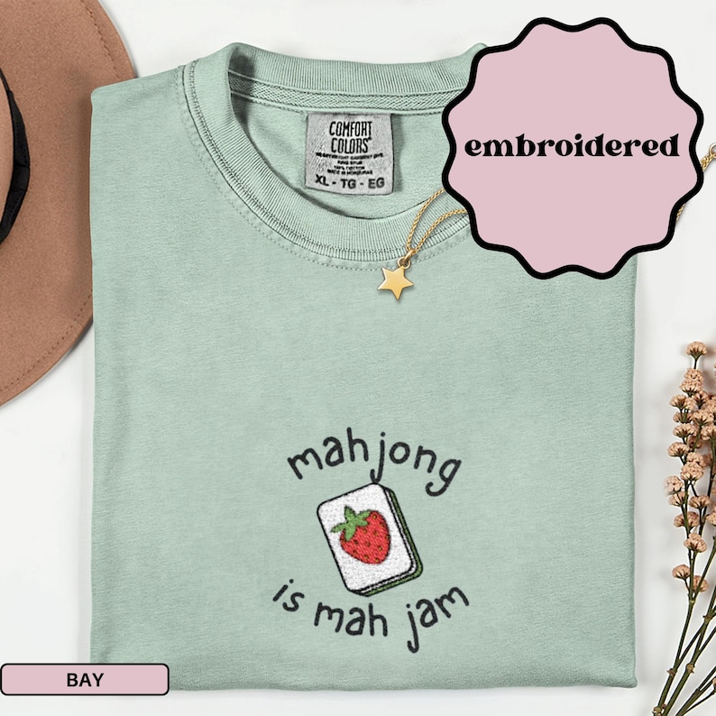 May include: A light green t-shirt with the text "mahjong is mah jam" and an embroidered strawberry tile design. The shirt is a Comfort Colors brand, size XL-TG-EG. A gold star necklace is also visible.