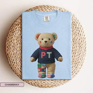 Peut inclure: T-shirt bleu clair avec un motif d'ours en peluche. L'ours porte un pull bleu marine avec les lettres "PT", une écharpe rouge et un pantalon patchwork. L'ours a des détails rouges, verts et bleus sur les oreilles et le visage. Le t-shirt est plié sur un support tressé.