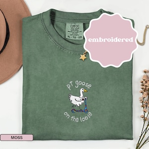 May include: Moss green Comfort Colors t-shirt with an embroidered design. The design features a white goose riding a scooter with the text "pt goose on the loose". The shirt is labeled "embroidered" and is size XL-TG-EG.