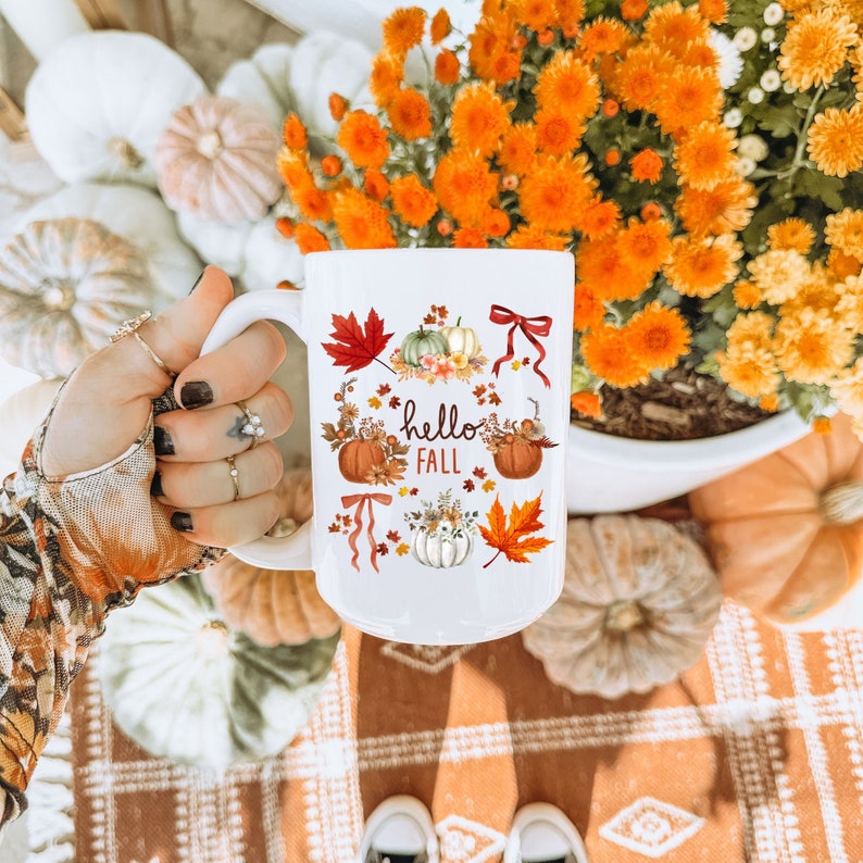 May include: White ceramic mug with a fall-themed design. The mug features a brown, orange, and white pumpkin, a red maple leaf, and the text "hello fall".