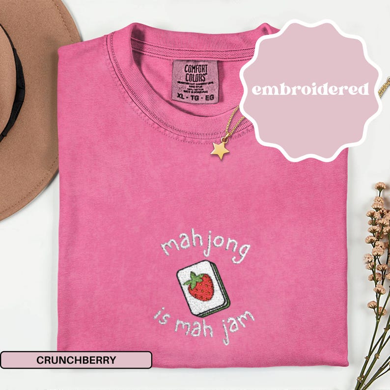 May include: A pink Comfort Colors t-shirt with the embroidered phrase "mahjong is mah jam" and a strawberry tile design. The shirt has a crew neck and short sleeves. A gold star pendant necklace is visible.