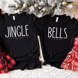 May include: Two black t-shirts with the words "Jingle" and "Bells" printed on them. The shirts are decorated with a string of colorful lights.