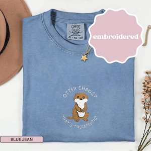 May include: A blue jean Comfort Colors t-shirt with an embroidered otter design. The otter is brown and white, with the words "Otter Chaos? That's Therapeutic!" in white. The shirt has a crew neck and short sleeves. A gold star necklace is visible.
