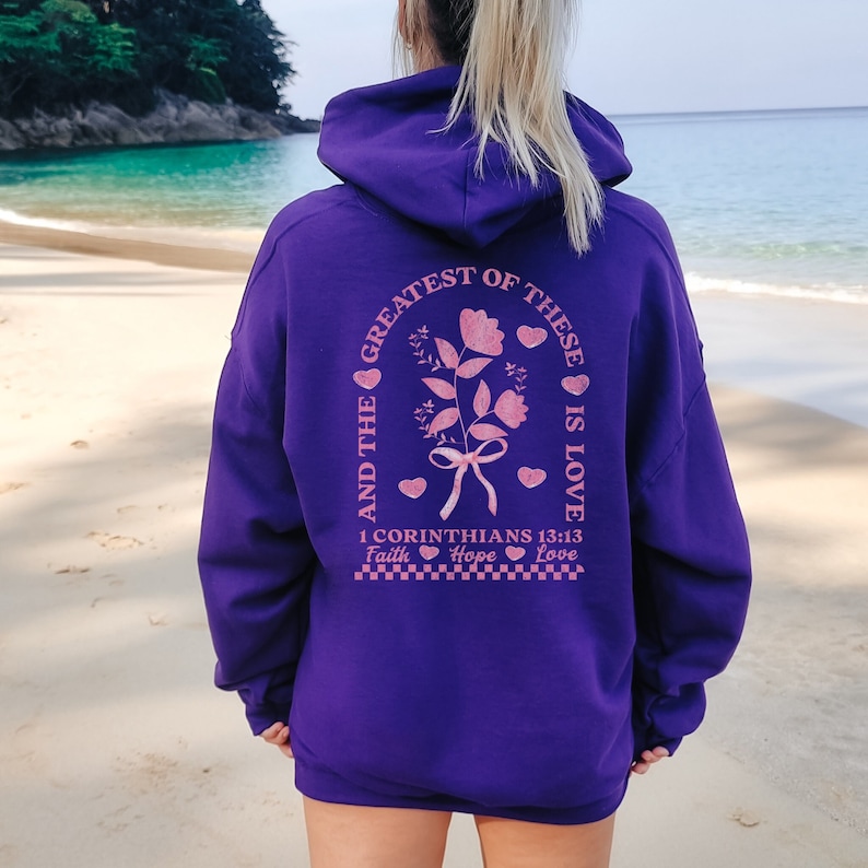 May include: Purple hoodie with a pink graphic on the back that says "And the greatest of these is love 1 Corinthians 13:13 Faith Hope Love" with a floral design and hearts.