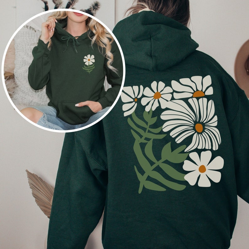 Puede incluir: Una sudadera con capucha verde oscuro con un dise&ntilde;o floral blanco en la espalda. Las flores son estilizadas y tienen un estilo retro.