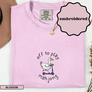 May include: A light pink, embroidered t-shirt with the words "off to play mahjong" and a cartoon duck riding a scooter. The shirt has a Comfort Colors tag and a gold star necklace.