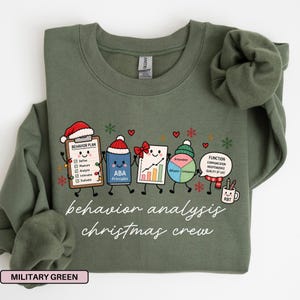 May include: A military green crewneck sweatshirt featuring a graphic of five cartoon characters representing behavior analysis tools wearing Christmas hats. The text reads behavior analysis christmas crew. The characters include a clipboard, a book, a chart, a pie graph, and a mug.