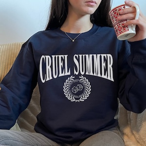 May include: A navy blue sweatshirt with the text "CRUEL SUMMER" in white, featuring a white wreath with dice and the text "DEVILS ROLL THE DICE, ANGELS ROLL THEIR EYES" in a circle.