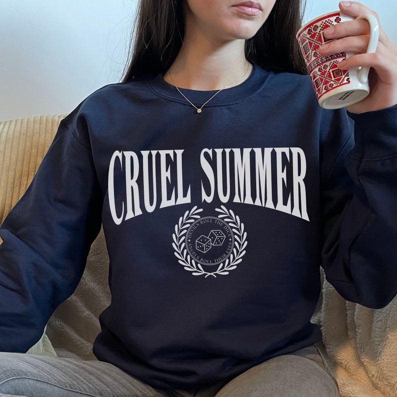 May include: A navy blue sweatshirt with the text "CRUEL SUMMER" in white, featuring a white wreath with dice and the text "DEVILS ROLL THE DICE, ANGELS ROLL THEIR EYES" in a circle.