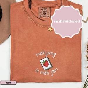 May include: A rust-colored t-shirt with the embroidered phrase "mahjong is mah jam" and a strawberry tile design. The shirt has a Comfort Colors tag and a size XL-TG-EG label.