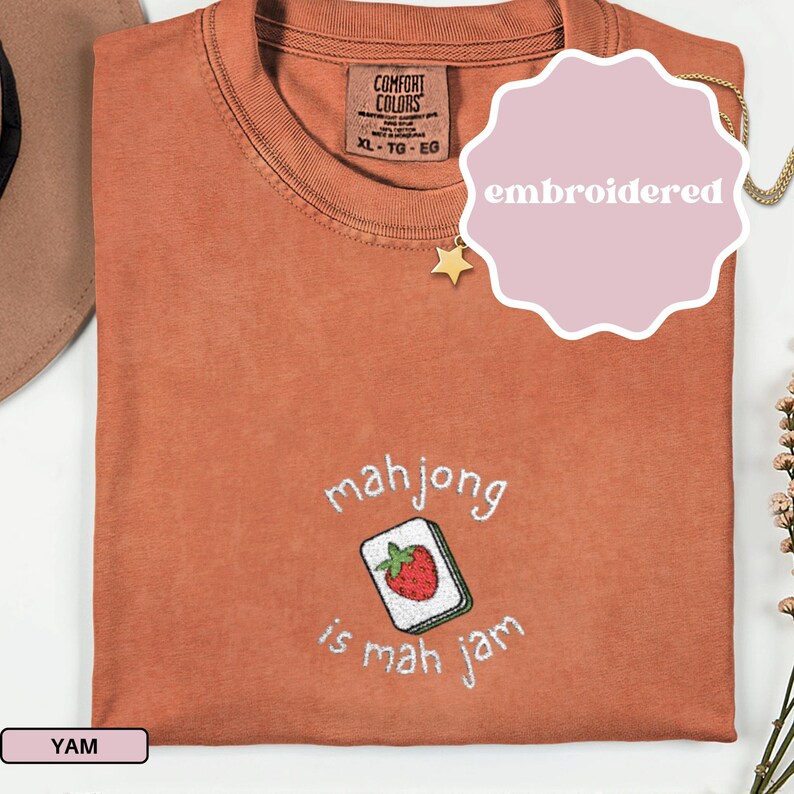May include: A rust-colored t-shirt with the embroidered phrase "mahjong is mah jam" and a strawberry tile design. The shirt has a Comfort Colors tag and a size XL-TG-EG label.