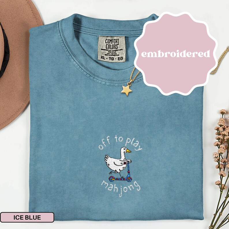 May include: Ice blue Comfort Colors t-shirt with an embroidered design of a white duck riding a scooter, with the text "off to play mahjong". A gold star pendant necklace is visible. The shirt is labeled "embroidered".