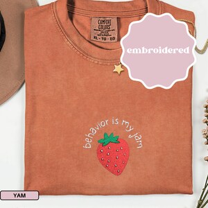 Peut inclure: Un t-shirt couleur rouille avec la phrase brod&eacute;e "behavior is my jam" entourant une fraise rouge avec des feuilles vertes. Le t-shirt a une &eacute;tiquette Comfort Colors et le mot "embroidered" sur un autocollant rose.