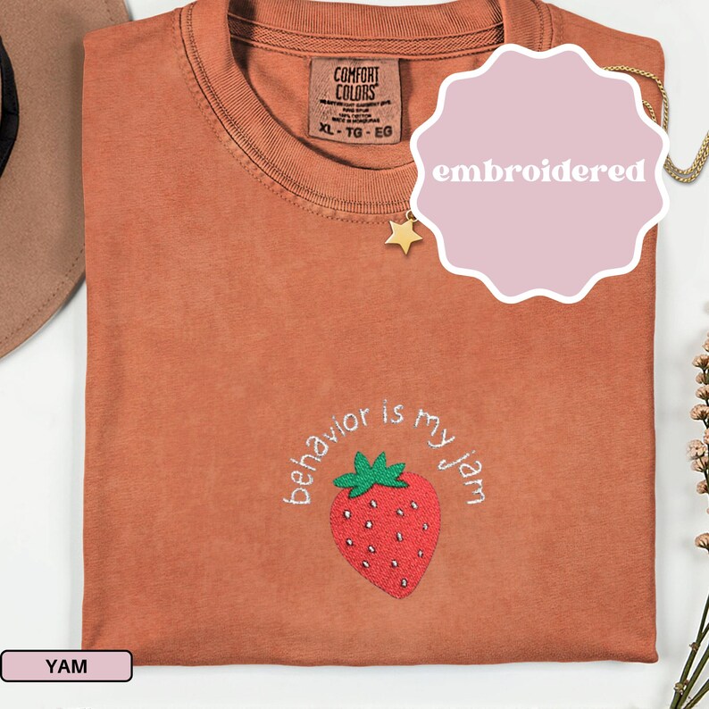 Peut inclure: Un t-shirt couleur rouille avec la phrase brod&eacute;e "behavior is my jam" entourant une fraise rouge avec des feuilles vertes. Le t-shirt a une &eacute;tiquette Comfort Colors et le mot "embroidered" sur un autocollant rose.