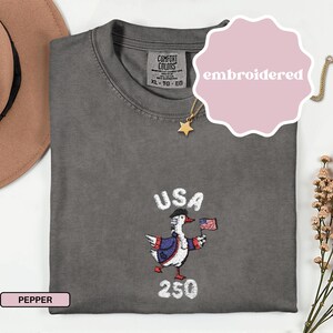 May include: A gray t-shirt with an embroidered design featuring a goose wearing a colonial hat and holding a small American flag. The word "USA" is above the goose, and "250" is below. The shirt has a tag that says "Comfort Colors".