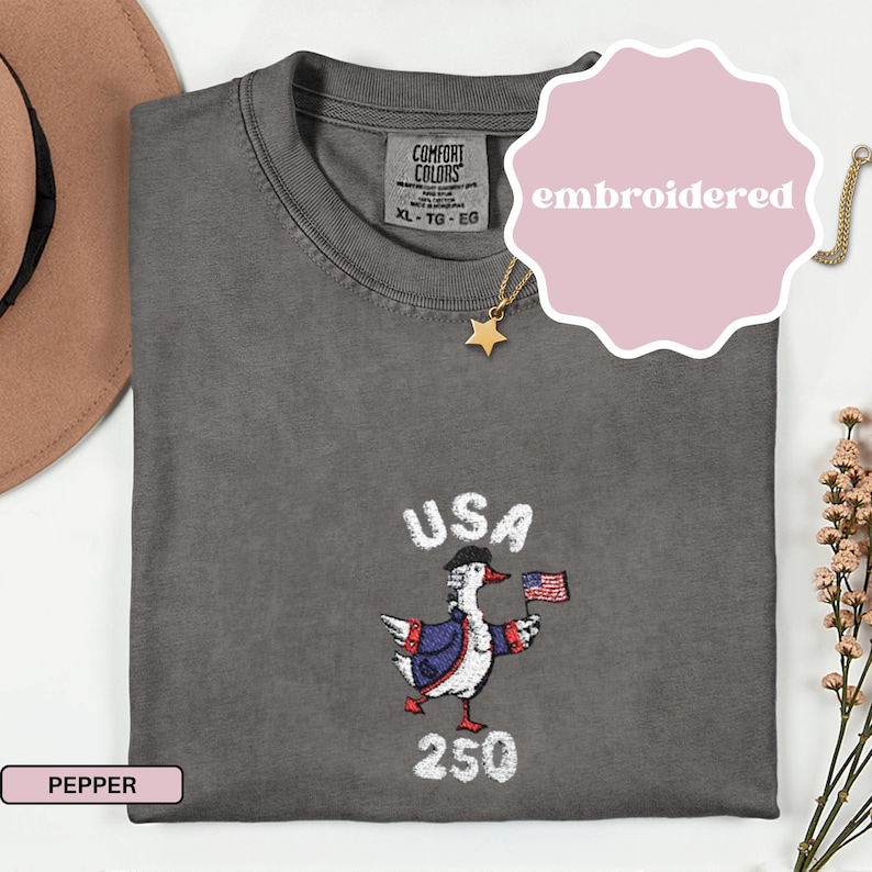 May include: A gray t-shirt with an embroidered design featuring a goose wearing a colonial hat and holding a small American flag. The word "USA" is above the goose, and "250" is below. The shirt has a tag that says "Comfort Colors".