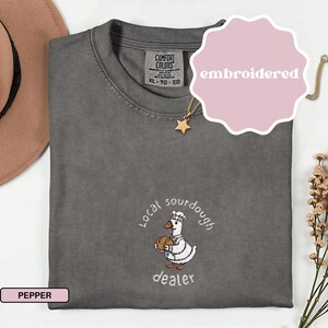 Puede incluir: Camiseta Comfort Colors color pimienta con un dise&ntilde;o bordado. El dise&ntilde;o presenta un pato de dibujos animados con gorro de chef y pan, con las palabras "Local sourdough dealer". Talla XL-TG-EG.