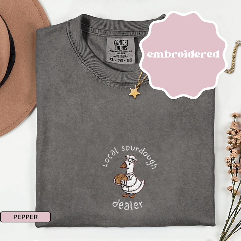 Puede incluir: Camiseta Comfort Colors color pimienta con un dise&ntilde;o bordado. El dise&ntilde;o presenta un pato de dibujos animados con gorro de chef y pan, con las palabras "Local sourdough dealer". Talla XL-TG-EG.