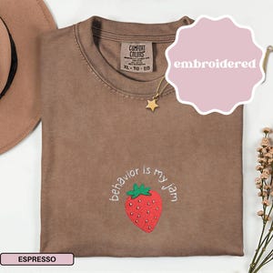 Peut inclure: T-shirt couleur expresso avec une fraise brod&eacute;e et l'inscription "behavior is my jam". Le t-shirt a un col rond et des manches courtes. Un collier pendentif &eacute;toile dor&eacute;e et un chapeau marron sont &eacute;galement visibles.