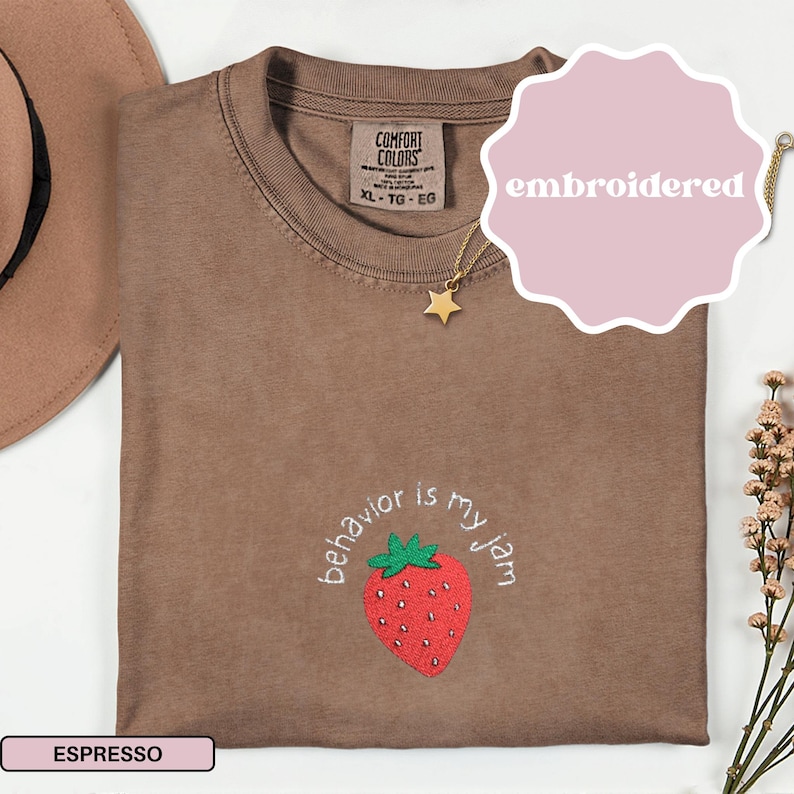 Peut inclure: T-shirt couleur expresso avec une fraise brod&eacute;e et l'inscription "behavior is my jam". Le t-shirt a un col rond et des manches courtes. Un collier pendentif &eacute;toile dor&eacute;e et un chapeau marron sont &eacute;galement visibles.