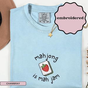 May include: Light blue chambray t-shirt with the text "mahjong is mah jam" and an embroidered mahjong tile with a red strawberry. The shirt has a crew neck and short sleeves. A gold star pendant necklace is also visible.