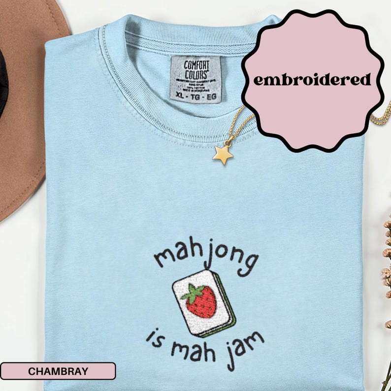 May include: Light blue chambray t-shirt with the text "mahjong is mah jam" and an embroidered mahjong tile with a red strawberry. The shirt has a crew neck and short sleeves. A gold star pendant necklace is also visible.