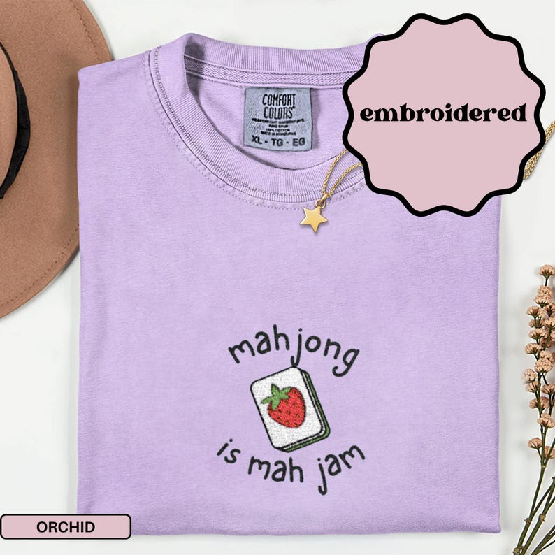 May include: Orchid-colored t-shirt with the embroidered text "mahjong is mah jam" and a strawberry-themed mahjong tile design. The shirt has a Comfort Colors tag and is size XL-TG-EG. A gold star pendant necklace is draped over the shirt.