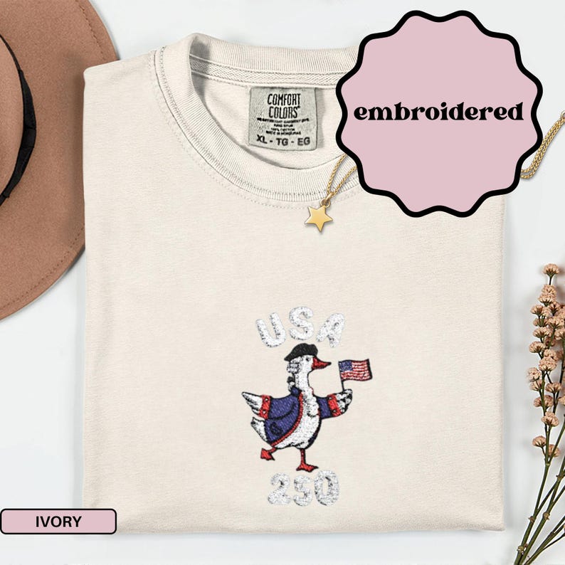 May include: Ivory-colored Comfort Colors t-shirt with an embroidered design. The design features a goose wearing a hat and jacket, holding a US flag, with the word "USA" above and "230" below. The shirt has a crew neck and a tag that reads "XL - TG - EG".