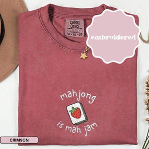 May include: Crimson-colored t-shirt with embroidered text and a mahjong tile design. The text reads "mahjong is mah jam" with a strawberry tile. The shirt has a Comfort Colors tag and a star pendant necklace.