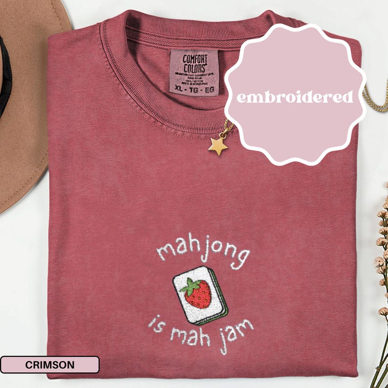 May include: Crimson-colored t-shirt with embroidered text and a mahjong tile design. The text reads "mahjong is mah jam" with a strawberry tile. The shirt has a Comfort Colors tag and a star pendant necklace.