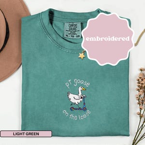 May include: A light green Comfort Colors t-shirt with an embroidered design of a goose riding a scooter. The text "pt goose on the loose" is below the graphic. The shirt is XL-TG-EG.
