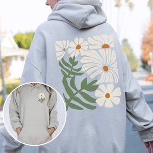 Puede incluir: Una sudadera con capucha gris claro con un dise&ntilde;o floral de margaritas blancas en la espalda. El dise&ntilde;o presenta un grupo de margaritas blancas con hojas verdes.