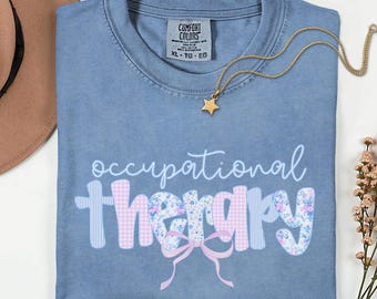 Op de afbeelding: Blauw spijkerbroekkleurig T-shirt met de woorden "occupational therapy" in een speels lettertype. Het woord "therapy" is versierd met een bloemen- en ruitpatroon. Een gouden sterrenketting ligt op het shirt.