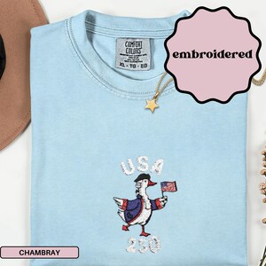 May include: Light blue embroidered t-shirt with a cartoon goose wearing a hat and holding a US flag. The word "USA" is above the goose, and the number "230" is below. The shirt is labeled "Chambray" and "embroidered".
