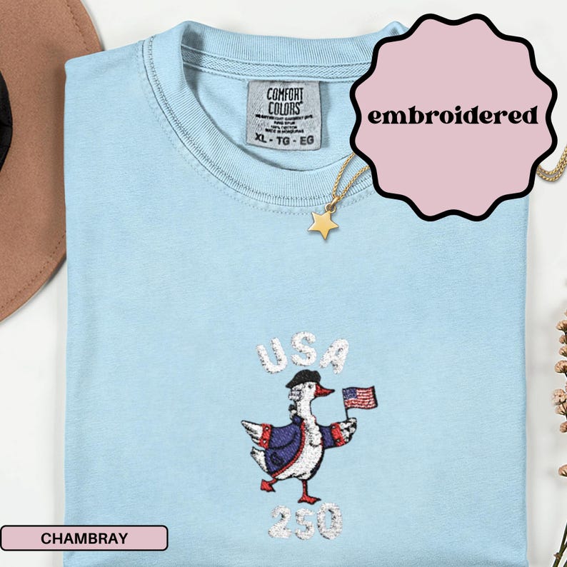May include: Light blue embroidered t-shirt with a cartoon goose wearing a hat and holding a US flag. The word "USA" is above the goose, and the number "230" is below. The shirt is labeled "Chambray" and "embroidered".