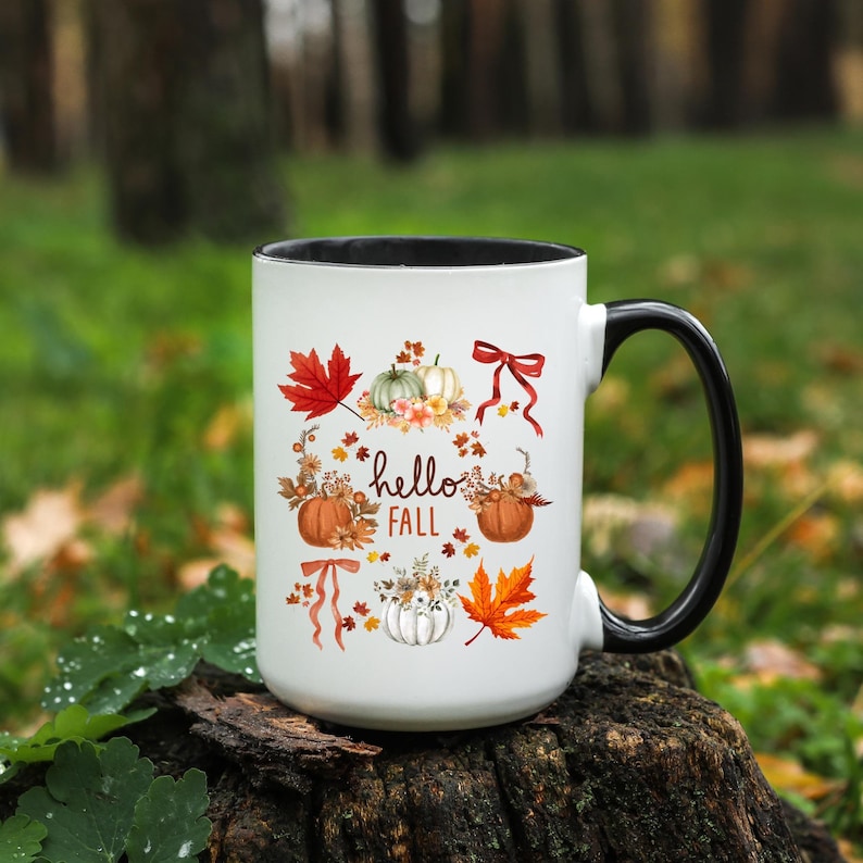 May include: White ceramic mug with a black rim and handle. The mug features a fall-themed design with pumpkins, leaves, and the text "hello fall".