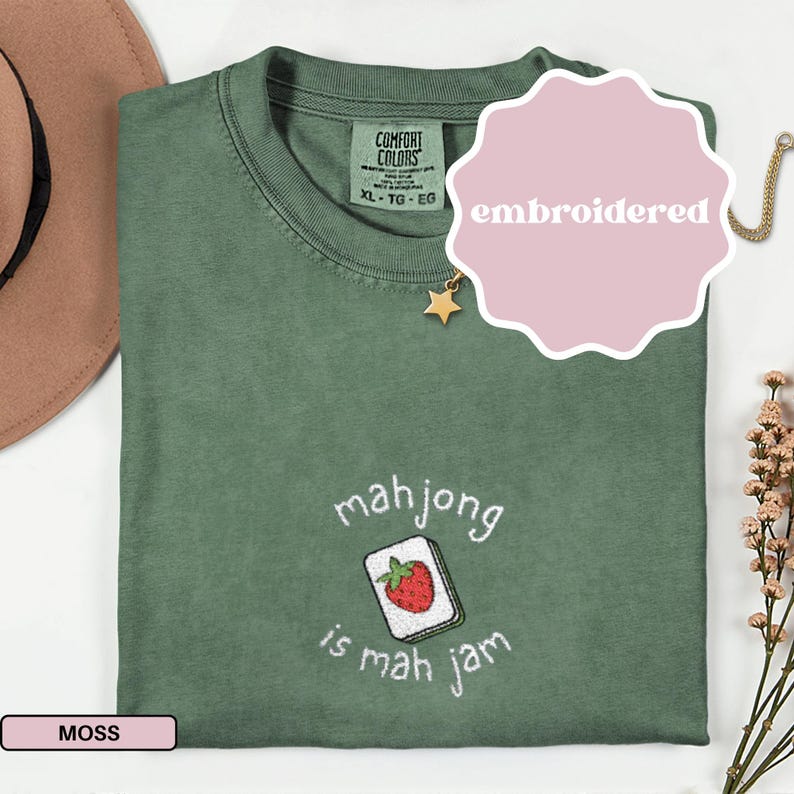 May include: Moss green t-shirt with an embroidered design. The design features a mahjong tile with a strawberry and the phrase "mahjong is mah jam". The shirt has a Comfort Colors tag and the word "embroidered" on a pink sticker.