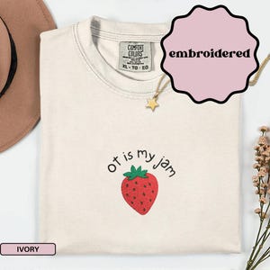 Può includere: T-shirt color avorio con la scritta ricamata "ot is my jam" sopra una fragola rossa. La maglietta ha un girocollo e maniche corte. Una collana d'oro con un ciondolo a stella è drappeggiata sulla maglietta.