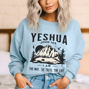 May include: Light blue sweatshirt with a graphic print of a mountain range, a bear, and birds. The text "YESHUA JOHN 14:6 I AM THE WAY. THE TRUTH. THE LIFE." is printed on the sweatshirt.