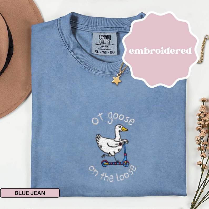 Może przedstawiać: Niebieska koszulka Comfort Colors z dżinsu z haftowanym wzorem. Wz&oacute;r przedstawia białą gęś jadącą na skuterze z napisem "of goose on the loose". Widoczny jest r&oacute;wnież złoty naszyjnik z gwiazdką i r&oacute;żowa metka z napisem "embroidered".