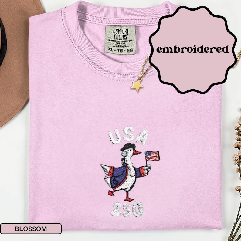 May include: Light pink Comfort Colors t-shirt with an embroidered design of a goose wearing a hat and holding a US flag. The word "USA" is above the goose, and "230" is below. The shirt has a crew neck and a tag that says "XL - TG - EG".