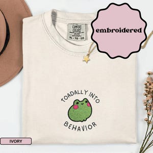 Puede incluir: Camiseta Comfort Colors color marfil con un diseño de rana bordada y la frase "TOADALLY INTO BEHAVIOR". La camiseta tiene cuello redondo y una etiqueta XL-TG-EG. Se ve un collar dorado con un colgante de estrella.
