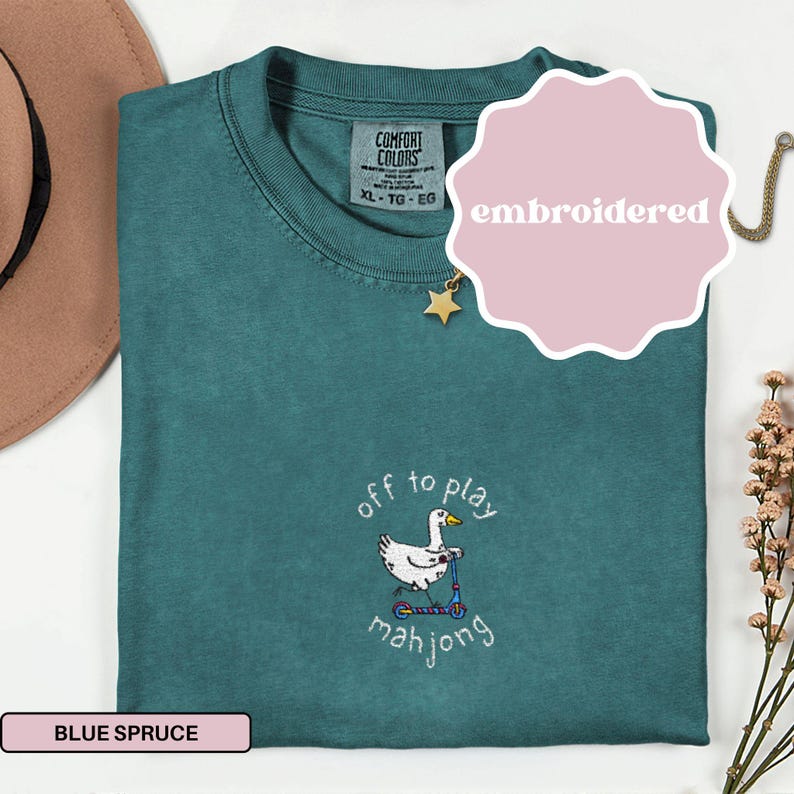 May include: Blue spruce Comfort Colors t-shirt with an embroidered design. The design features a white duck riding a scooter with the words "off to play mahjong". The shirt is labeled XL-TG-EG.
