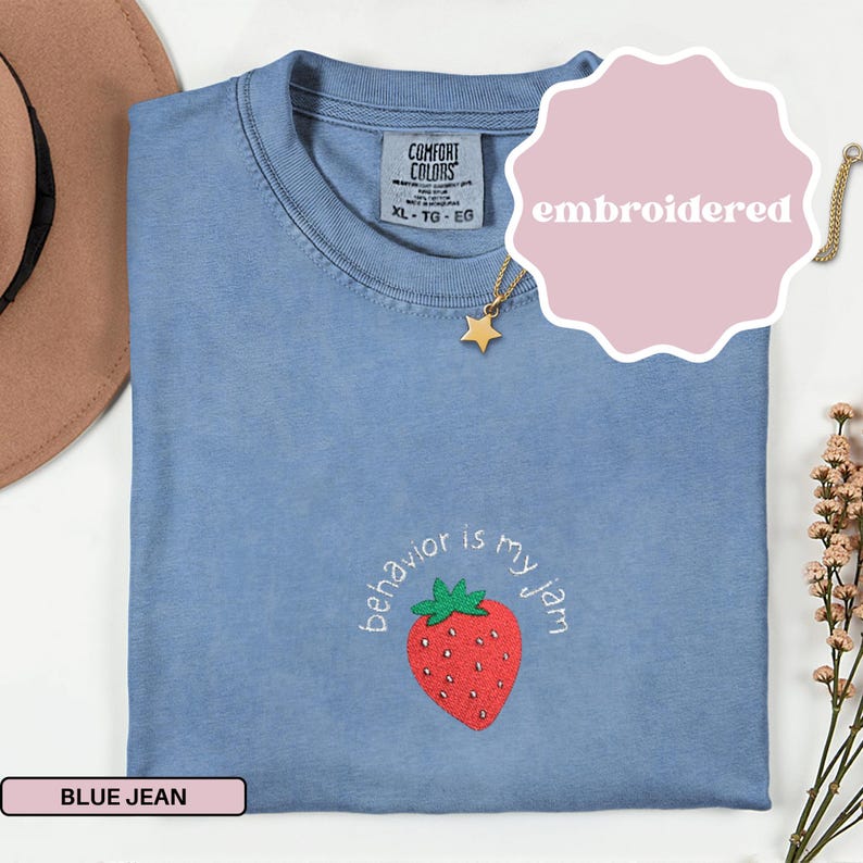 Peut inclure: T-shirt bleu jean Comfort Colors avec une fraise brod&eacute;e et la phrase "behavior is my jam". Le t-shirt a un col rond et des manches courtes. Un collier pendentif &eacute;toile dor&eacute;e est &eacute;galement visible.