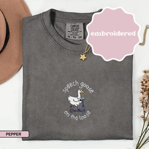 May include: Pepper-colored t-shirt with an embroidered design featuring a goose riding a scooter, with the text "speech goose on the loose". The shirt has a crew neck and short sleeves. A pink sticker with the word "embroidered" is on the top right.