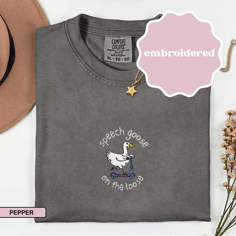 May include: Pepper-colored t-shirt with an embroidered design featuring a goose riding a scooter, with the text "speech goose on the loose". The shirt has a crew neck and short sleeves. A pink sticker with the word "embroidered" is on the top right.