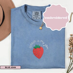 May include: A blue jean Comfort Colors t-shirt with an embroidered strawberry and the phrase "speech is my jam." The shirt has a crew neck and short sleeves. A gold star pendant necklace is visible.