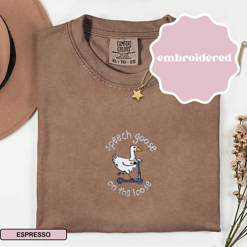 May include: Espresso-colored t-shirt with an embroidered design of a goose on a scooter, with the text "speech goose on the loose". The shirt has a Comfort Colors tag and a star pendant necklace.